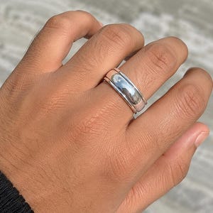 Könnte beinhalten: Ein polierter silberfarbener Ring, bestehend aus einer breiten Schiene mit glatter, reflektierender Oberfläche und einem dünneren, separaten silberfarbenen Ring, der darüber liegt und einen gestapelten Look erzeugt.