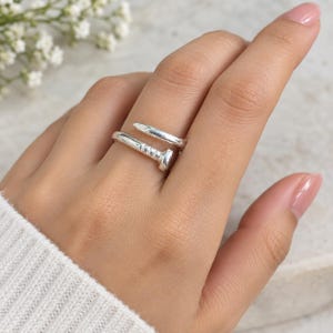 May include: A polished silver ring shaped like a bent nail that wraps around the finger. The design features a flat, circular nail head on one end and a tapered, pointed tip on the other, with a smooth, reflective metallic finish throughout the band.