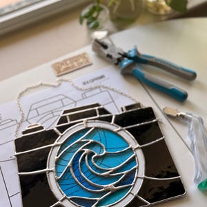 May include: A stained glass camera with a wave design in shades of blue and teal. The camera is black with a silver chain for hanging. The artist's signature, "Nicki Evans Photo," is visible.