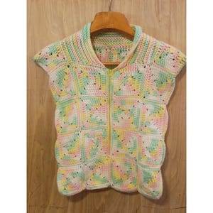 May include: A sleeveless crochet waistcoat constructed from square motifs in pastel shades of pink, yellow, green, and white. The garment features a central yellow zip closure extending from the collar to the hem, with a high neckline and textured yarn.