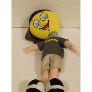 May include: A plush doll with a large yellow circular head, a black baseball cap, and a gray neck spring. The doll wears a dark gray t-shirt with the word BEAMER printed in yellow, tan shorts, and black and white shoes with a rounded design.