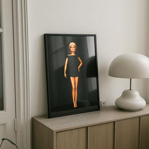 Puede incluir: Impresión artística enmarcada que presenta una muñeca Barbie con un vestido negro. La muñeca tiene el pelo rubio y posa sobre un fondo oscuro. La impresión se exhibe en un mueble de madera, junto a una lámpara blanca.