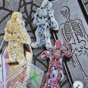 Könnte beinhalten: Eine Sammlung von dekorativen Gegenständen zum Thema Skelett. Das Bild zeigt mehrere Skelettfiguren in Gold-, Silber- und Rottönen sowie eine Skelettillustration. Die Objekte sind vor einem strukturierten Hintergrund platziert.