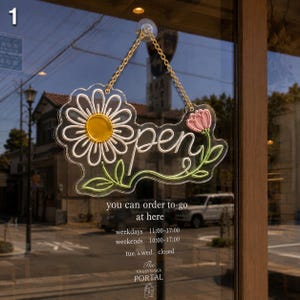 Op de afbeelding: Een hangend bord van helder acryl met het woord open in witte letters, voorzien van een geel-witte margriet aan de linkerkant en een roze bloem met groene stelen aan de rechterkant. Het bord hangt aan een gouden ketting aan een raam.