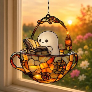 Puede incluir: Un atrapasueños de vidrieras con forma de taza de té con un asa a cada lado, con piezas de vidrio naranja, amarillo y marrón. Dentro de la taza hay una pequeña figura de fantasma blanco que sostiene un libro abierto. Un asa arqueada cuelga arriba.