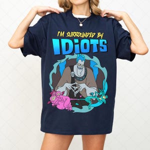 May include: Dark blue t-shirt with a cartoon graphic. The design features the text "I'M SURROUNDED BY IDIOTS" above an illustration of Hades and two creatures. The graphic uses blue, pink, and grey colours.