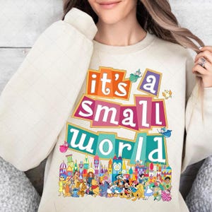 Puede incluir: Sudadera color crema con la frase "it's a small world" en texto colorido y superpuesto. El diseño incluye una ilustración vibrante de varios personajes de dibujos animados y edificios, creando una estética caprichosa y alegre.