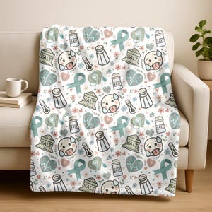 May include: White throw blanket with a repeating pattern of cartoon faces, salt shakers, thermometers, hearts, flowers, and teal ribbons. The word "SALT" is printed on some of the objects. The blanket is draped over a white sofa.