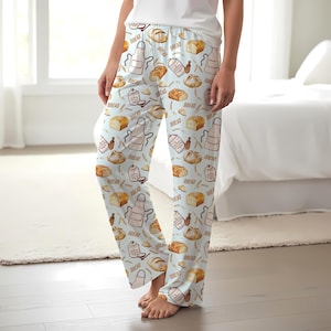 Pode incluir: Calças de pijama azul-claras com uma estampa de pães castanhos, aventais brancos, recipientes de farinha e a palavra BREAD em texto castanho. As calças têm um corte descontraído com cintura elástica e pernas direitas.