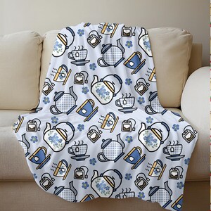 May include: A white throw blanket with a repeating pattern of teapots, teacups, tea bags, and blue flowers. The teapots and teacups are in blue and white, with yellow accents. The blanket is draped over a beige sofa.