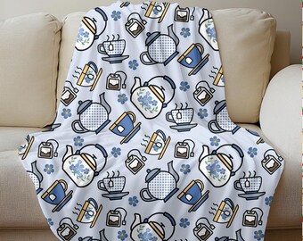 May include: A white throw blanket with a repeating pattern of teapots, teacups, tea bags, and blue flowers. The teapots and teacups are in blue and white, with yellow accents. The blanket is draped over a beige sofa.