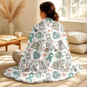 May include: A white blanket with a repeating pattern of salt shakers, thermometers, teal ribbons, hearts, and cartoon faces. The blanket is draped over a person sitting on the floor.