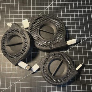 Puede incluir: Tres organizadores de cables negros y circulares con un diseño perforado y una barra central. Cada organizador tiene un cable USB-C blanco que se extiende desde él. Los artículos están dispuestos sobre una superficie de cuadrícula negra.