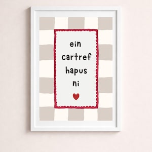 May include: A framed wall art print featuring a light beige and white checkered background. Centered is a white rectangular panel with a scalloped dark red border containing the black text "ein cartref hapus ni" and a small red heart icon.