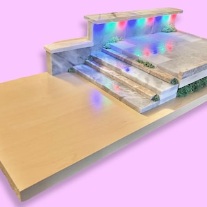 May include: A miniature stage model with a light wood base and marble accents. The stage features steps, a platform, and LED lights in blue, green, and red. Green foliage adds a natural touch to the design.