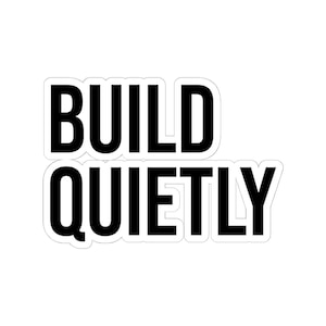 May include: A white sticker with a black border and the words "BUILD QUIETLY" in bold, black capital letters. The sticker has a clean, minimalist design.