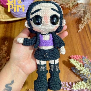 Puede incluir: Una muñeca de ganchillo hecha a mano con pelo negro, chaqueta negra, top corto morado, falda negra con un detalle de anillo plateado y botas negras con detalles de red en las piernas. La muñeca tiene ojos negros grandes, gargantilla y aros plateados.
