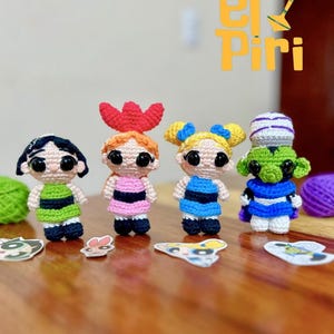May include: Four handmade crochet dolls, each with large black eyes and unique outfits. The dolls are in green, pink, and blue dresses, with matching hair accessories. The dolls are arranged on a wooden surface with small character cutouts.