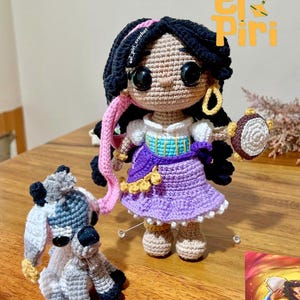 Puede incluir: Muñeca de ganchillo de una mujer con cabello negro, un vestido morado y una pandereta. Una figura de animal de ganchillo está al lado de la muñeca. El texto "El Piri" está en la esquina superior derecha.