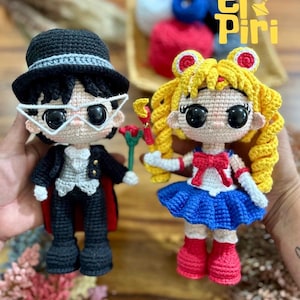 Puede incluir: Dos muñecos de ganchillo hechos a mano, uno vestido con un esmoquin negro con sombrero de copa y gafas, el otro con un uniforme de marinero con falda azul y pelo amarillo. Los muñecos sostienen accesorios.