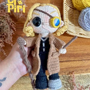 May include: A crochet doll with blonde hair, wearing a tan coat over a black outfit. The doll features a black eye patch over one eye, a blue and white monocle over the other, and holds a brown textured staff and a thin brown wand in its hands.