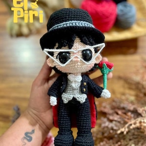 Könnte beinhalten: Gehäkelte Puppe eines Charakters in einem schwarzen Anzug, Zylinder und Brille, die eine rote Rose hält. Die Puppe hat einen roten Umhang und eine graue Weste. Der Text "El Piri" ist im Bild zu sehen.