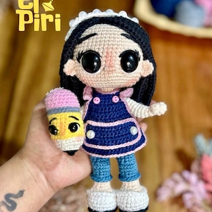 Puede incluir: Muñeca de ganchillo con cabello negro, ojos negros grandes y una diadema blanca. La muñeca lleva un vestido azul con ribete rosa y vaqueros azules. Sostiene un lápiz de ganchillo con una goma de borrar rosa. El texto "el Piri" está arriba a la izquierda.