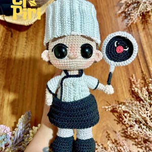 Puede incluir: Una muñeca de ganchillo hecha a mano vestida de chef, con un gorro blanco, una chaqueta blanca de doble botonadura y una falda negra. La muñeca tiene grandes ojos negros y sostiene una pequeña sartén negra con el centro rojo en una mano.