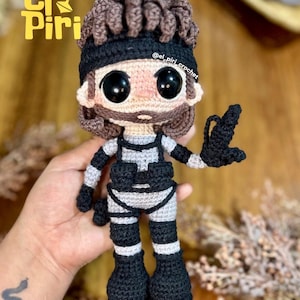 Puede incluir: Una muñeca de ganchillo que representa un personaje con pelo castaño, una cinta negra y barba. La figura lleva un traje táctico gris claro con correas negras, botas negras y guantes negros, sosteniendo un pequeño objeto negro en la mano.