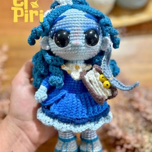 May include: A handmade crochet doll with blue hair and a blue dress. The doll has large black eyes and is holding a small book. The doll's hair is styled in curls, and it has a white collar. The doll is wearing blue shoes. The text "el Piri" is in the upper left corner.