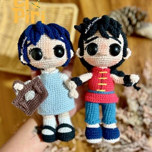 Puede incluir: Dos muñecas de ganchillo hechas a mano. Una muñeca viste un vestido azul claro y sostiene un libro marrón. La otra muñeca lleva una parte superior roja y pantalones azules. Ambas muñecas tienen grandes ojos negros y cabello oscuro. El texto "El Piri" y "el_piri_crochet" son visibles.