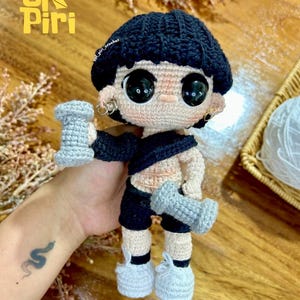 Puede incluir: Muñeca de ganchillo con pelo negro, pantalones cortos negros y zapatillas blancas, sosteniendo dos mancuernas grises. La muñeca tiene ojos negros grandes y lleva un top negro. Diseño único.