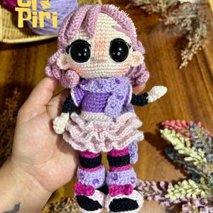 Puede incluir: Una muñeca de ganchillo con cabello morado claro, ojos negros grandes y cara beige. La muñeca viste una blusa morada con bufanda a juego, una falda rosa claro con volantes, calentadores a rayas moradas y negras, y zapatos con gemas.
