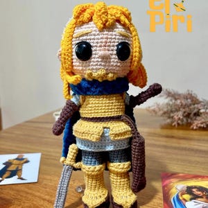 Puede incluir: Muñeca de ganchillo hecha a mano de un guerrero, con cabello amarillo, una capa azul y ropa tipo armadura en tonos amarillos y marrones. La muñeca sostiene una espada y una bolsa marrón. El texto "El Piri" es visible en el fondo.