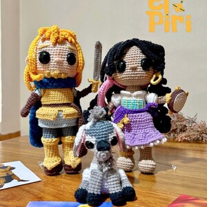 Puede incluir: Muñecos de ganchillo de un guerrero, una mujer y un burro, con una espada y una pandereta. El guerrero lleva un atuendo amarillo y azul, la mujer un vestido morado, y el burro es gris y blanco. Los muñecos están sobre una superficie de madera.