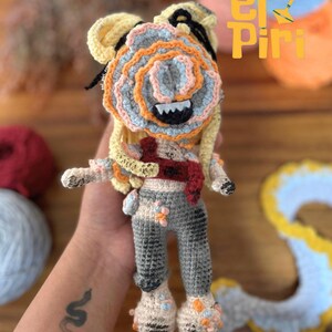 Könnte beinhalten: Handgefertigte Häkelpuppe mit einzigartigem Design. Die Puppe hat einen mehrschichtigen Kopf in Gelb-, Blau-, Orange- und Rosatönen mit einem grinsenden Mund. Sie trägt eine graue Hose, eine rote Schärpe und florale Akzente. Das Design der Puppe enthält den Text "El Piri".