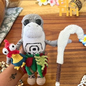 May include: Crocheted shark doll with a white belly, gray body, and black eyes. The doll is holding a crocheted rooster and a white staff with a brown handle. The shark is wearing a crocheted skirt with red, green, and brown accents.