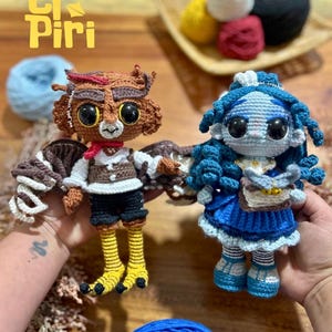 Peut inclure: Deux poupées au crochet faites à la main. L'une est un hibou brun avec de grands yeux, portant un gilet et un short. L'autre est une poupée bleue avec des cheveux bleus et une robe bleue. Les poupées sont exposées avec du fil et le texte "El Piri".
