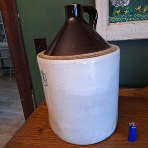 May include: A large, antique stoneware jug with a dark brown, cone-shaped top and a white, cylindrical body. The jug has a handle and a small blue emblem. A blue lighter is next to the jug. The jug is likely used for storing liquids.