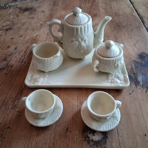 May include: A miniature off-white ceramic tea set featuring a teapot with a lid, a sugar bowl with a lid, a creamer, two teacups with matching saucers, and a rectangular serving tray, all decorated with embossed floral and lattice patterns.