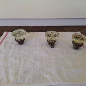 May include: Three antique glass doorknobs with a faceted design, each mounted on a dark metal base. The knobs are clear with a slight yellow tint, resting on a white cloth with a red border. The background is a wooden surface.