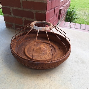 May include: A rusty, round metal bird feeder with a wire cage. The feeder has a circular base and a cage that extends upwards. The metal has a weathered, aged appearance, suggesting outdoor use. The feeder sits on a concrete surface.