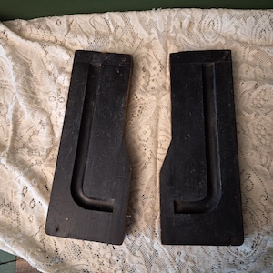 May include: Two antique, black wooden molds with a curved, U-shaped design. The molds are placed on a white lace fabric, suggesting a vintage or craft theme. The wood has a worn appearance.