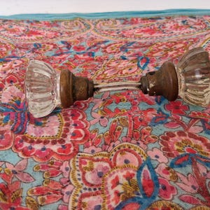 May include: A vintage door handle set featuring two clear, fluted glass knobs attached to a central metal spindle. The knobs have a distinct ribbed texture and are connected by a weathered, dark metallic base that shows signs of age and patina.