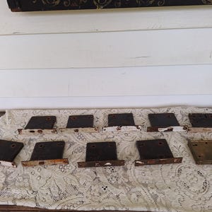 May include: A collection of antique metal door latches. The latches are rectangular with a dark, aged finish and white or rust-colored accents. They are arranged on a lace tablecloth.