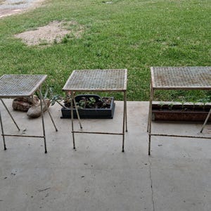 May include: Three vintage metal side tables with square, mesh tops and slender, angled legs. The tables are painted in a faded off-white color, showing signs of age and wear. They are arranged on a concrete surface with a grassy background.