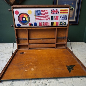 May include: A vintage wooden desk with an open lid, revealing compartments and drawers. The desk features a color wheel chart and flags of various countries. A black bird is painted on the desk's surface.