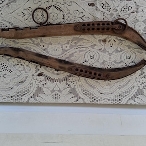 May include: Two antique wooden ox yokes, featuring a curved design with multiple holes and metal ring attachments. The yokes are a weathered brown color, suggesting age and use. They are displayed on a white lace background.