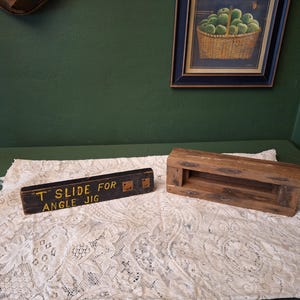 May include: Two wooden woodworking jigs sit on a white lace cloth. One is a rectangular block with a slot, the other is a black block with the text "T SLIDE FOR ANGLE JIG" in gold lettering. A painting of apples hangs in the background.