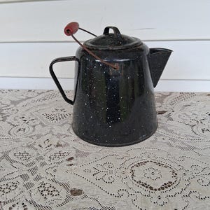 May include: A black enamel coffee pot with a white speckled pattern, featuring a metal handle with a red wooden grip, a matching lid, and a side spout. The pot has a dark, weathered finish with visible signs of age and use on the metal surfaces.